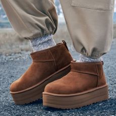 A close up of someone wearing some beige trousers, grey socks and some brown UGG-style slipper boots.