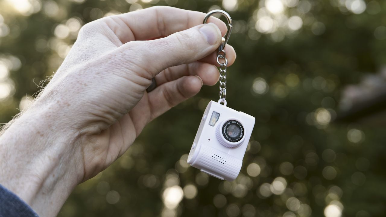 Miniature pink keychain camera, the 'Photo Creator Mini Digital Camera series 3', in user's hand, dangling from keychain