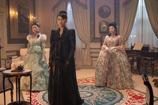 Three women wearing Regency gowns (Michelle Mao as Rosamund Li, Katie Leung as Lady Araminta Gun, and Isabelle Wei as Posy Li) stand in a drawing room, in 'Bridgerton' season 4.