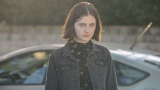 Miranda Frangou in a black denim jacket and flowered top standing in front of a car as Lorelei in The Secrets She Keeps.