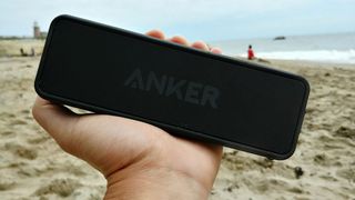 A Soundcore 2 speaker held in an outstretched hand on a beach