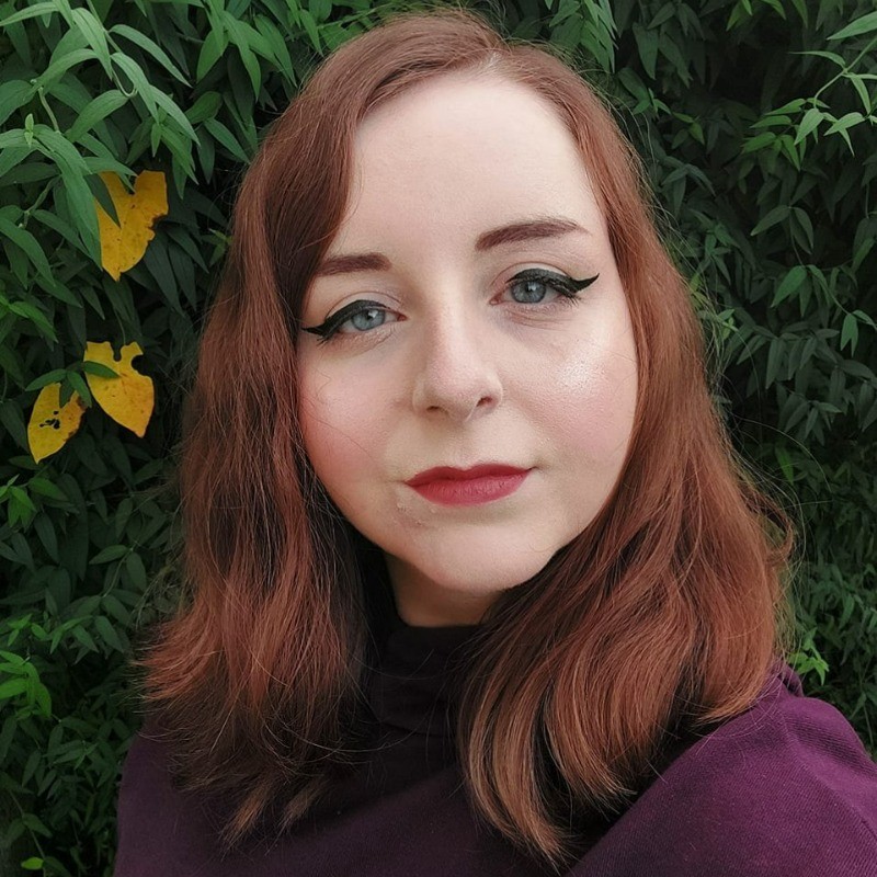 Jess Kinghorn stands in front of a hedge with lots of green leaves, and three prominent yellow leaves in the frame. This is a profile image and she isn't smiling.