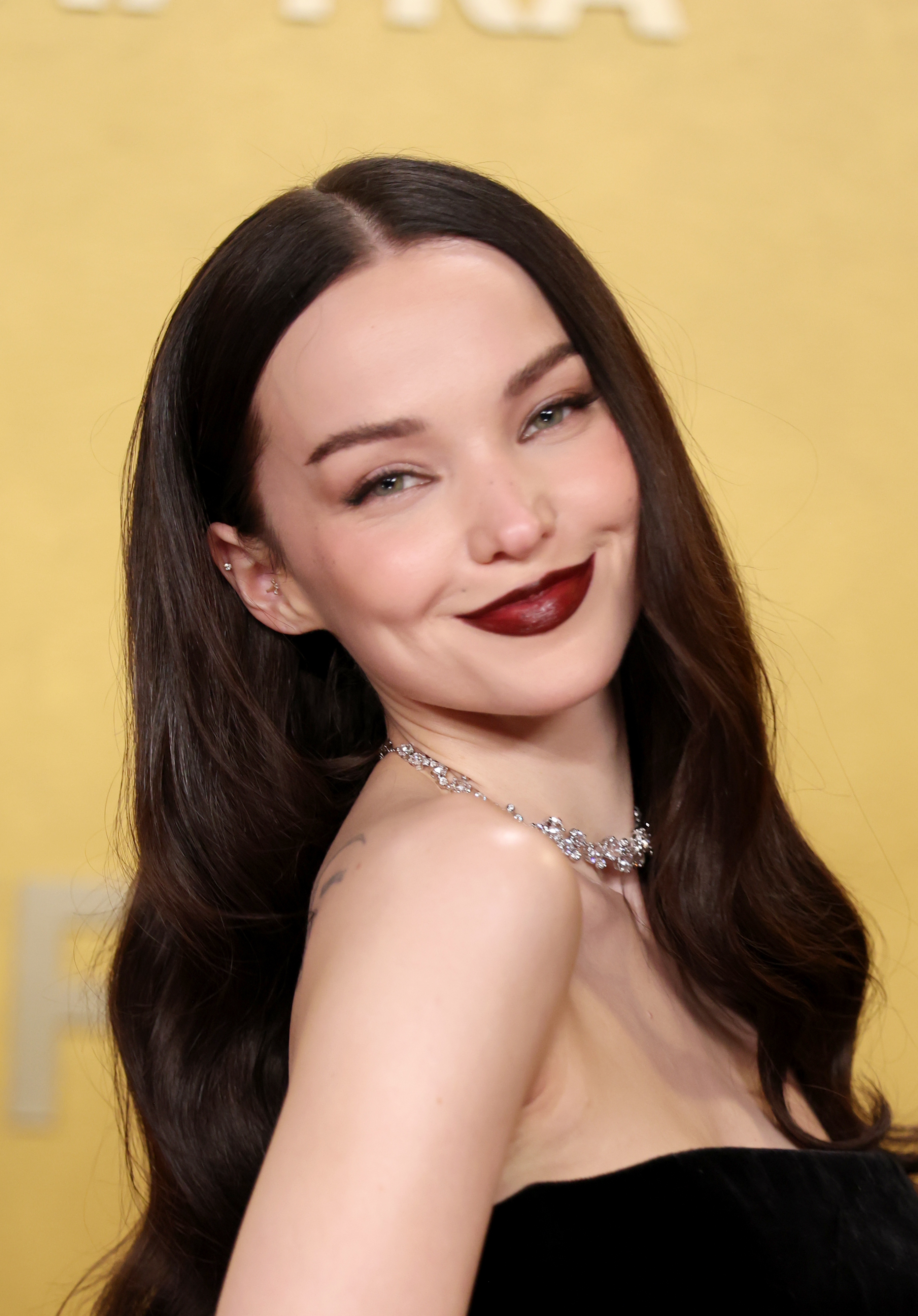 a young white woman with long, dark hair wears dark red lipstick and poses in front of a yellow backdrop on a red carpet