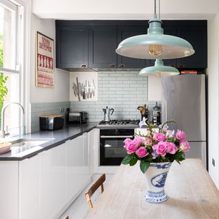 An L-shaped kitchen with black and white cabinets, a white brick splashback and two mint green pendant lights hanging over a wooden kitchen table