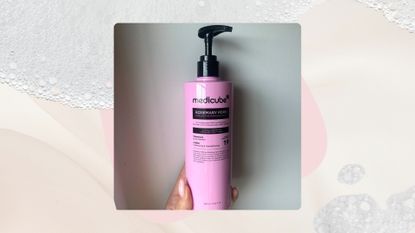Image of hand holding pink bottle of Medicube's Rosemary PDRN Shampoo on a pale pink background with foam details