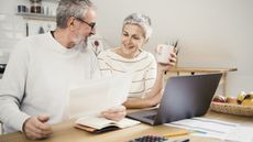 A retired couple smile as they look over paperwork at a table while a laptop is open in front of them.