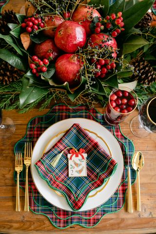 Christmas table setting on a wooden dining table. There is a tartan placemat with a white plate and a matching napkin on top of the plate. The centerpiece is made out of magnolia leaves and pomegranates