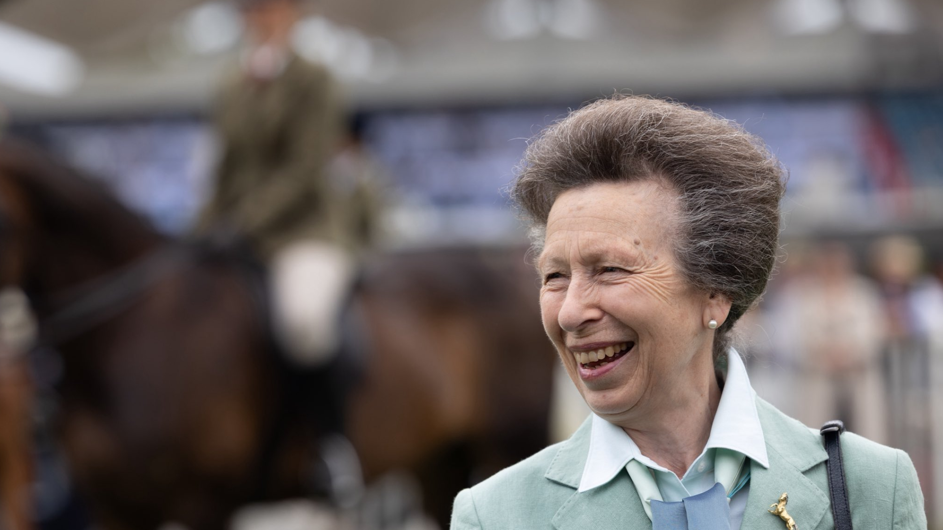 Princess Anne wearing a mint green blazer with a horse rider in the background
