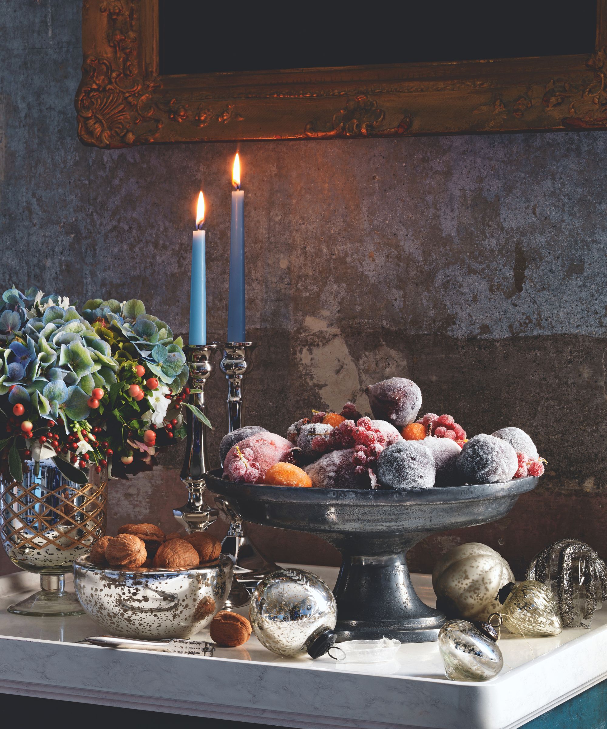 Silver and iron vases filled with figs and berries, next to blue taper candles and silver baubles