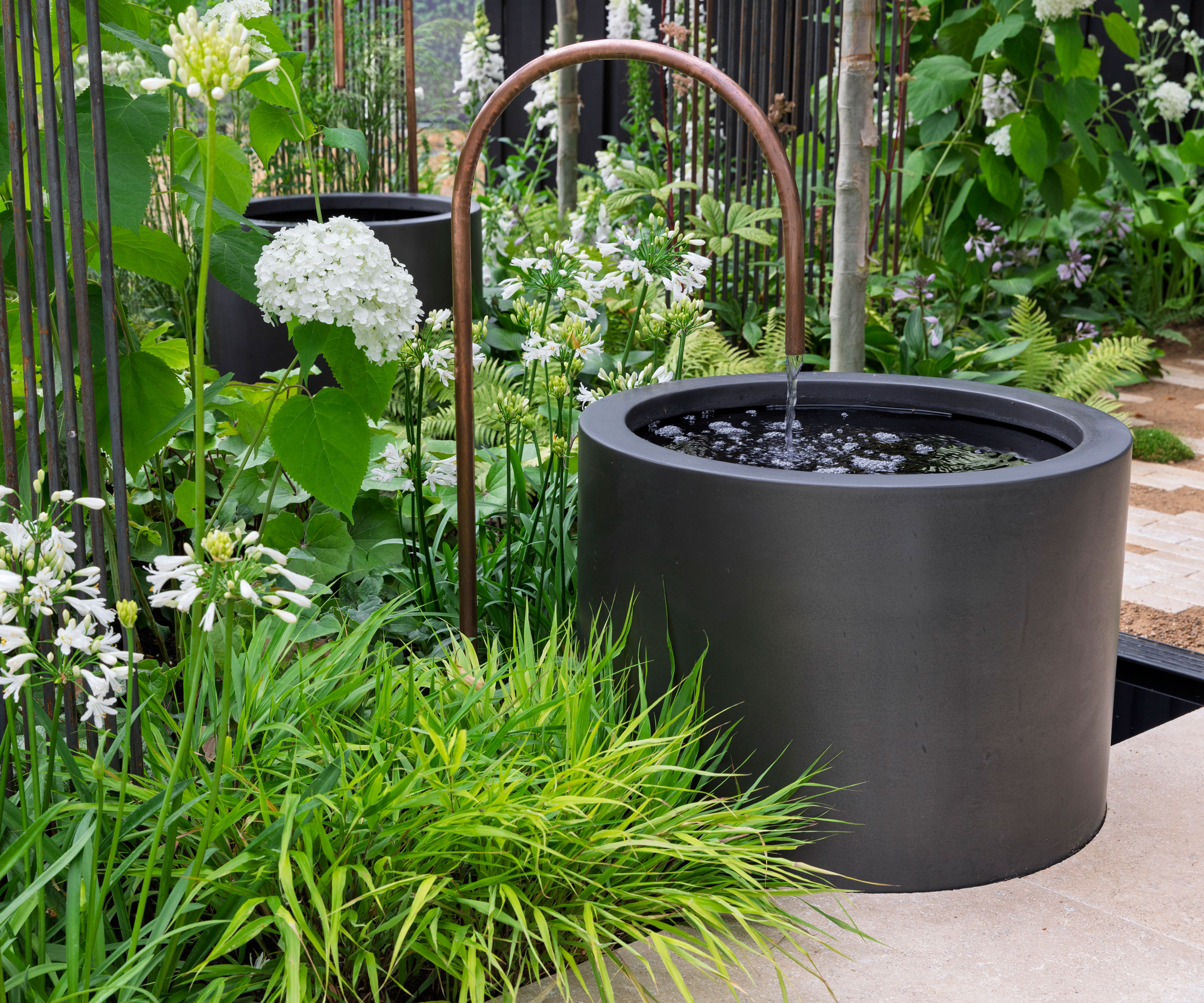 two small black water features with copper spouts in paved garden with planting including ornamental grasses and hydrangeas