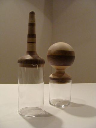 Two cynlinder glass bottles in different heights with tops made from different shades of wood photographers on a grey surface with a brown background