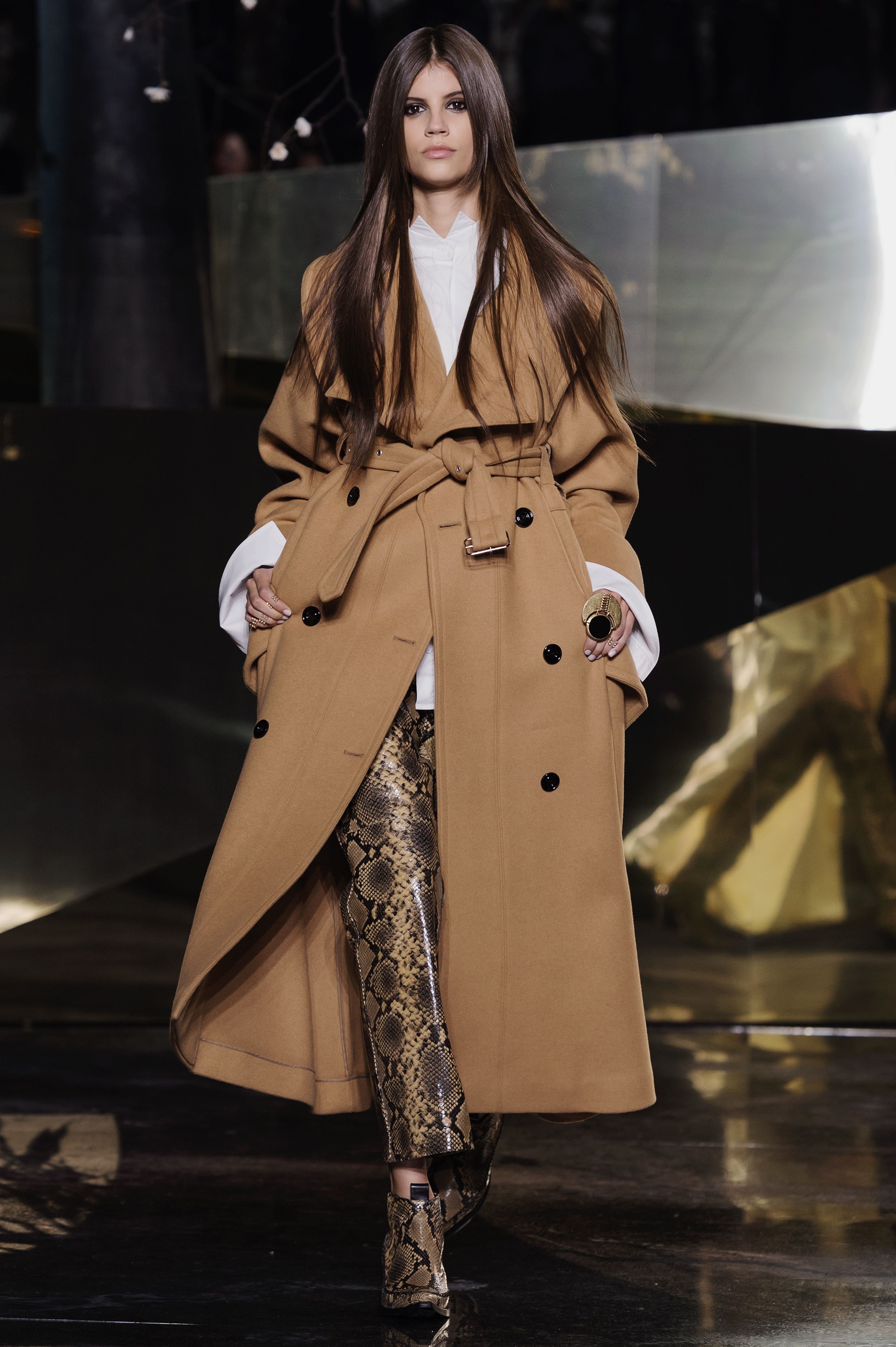 A model walks the runway wearing a coat and snake print trousers and boots.