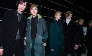 Four male and female models wearing looks from Costume National's collection. The first male model is wearing a black patterned suit and waistcoat with a grey shirt. Next to him is a female model wearing a black shirt, dark green trousers and a blue panelled coat with off-white coloured fur. The second male mode is wearing a white turtle neck and black coat. And beside him is a female model wearing a black top and dark green coat