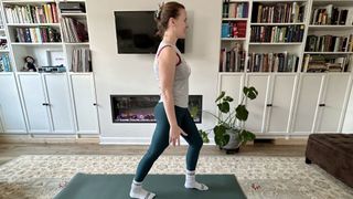 Woman exercising in domestic setting. She is wearing green leggings and sleeveless gray top. She is standing with one foot slightly behind her and one foot slightly in front of her.