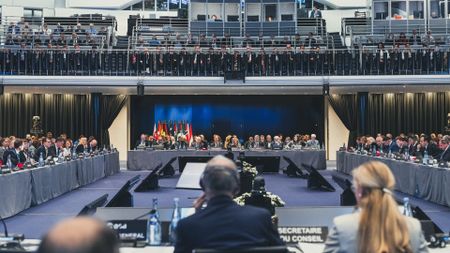 A series of people sit at four different tables making a square to face each other with various flags of their corresponding countries next to them.