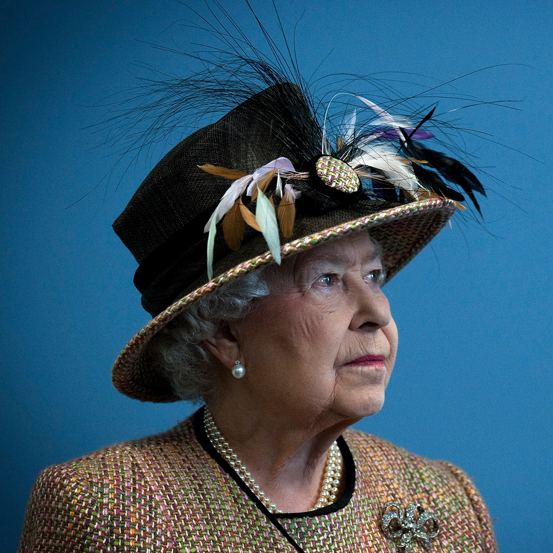 Queen Elizabeth wearing a feathered hat and tan jacket standing in front of a blue wall
