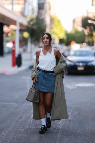 woman in mini skirt outfit of denim skirt, white tank top, trench coat, loafers
