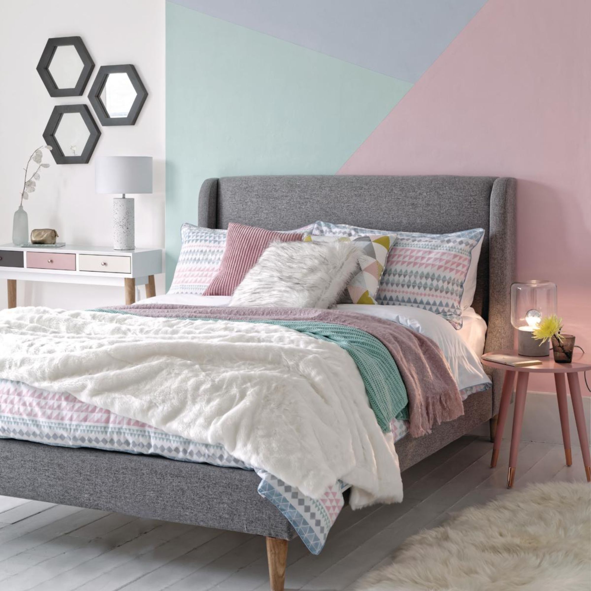 Double bed with multicoloured cushions and throws on a white wooden floor with a geometric design on the wall behind.