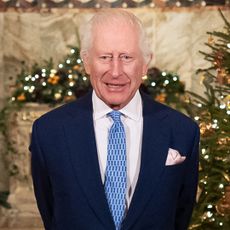 King Charles wearing a blue suit and tie standing in front of a lit Christmas tree 