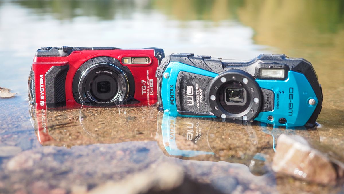 OM System Tough TG-7 and Pentax WG-90 cameras sitting in water, with a view of trees reflected in the water behind them