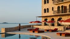 A sunset scene caught on the outdoor patio and pool of a hotel, decked out with dark red parasols and matching reclining loungers, an infinity pool, and uninterrupted sea views