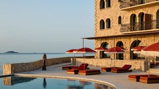 A sunset scene caught on the outdoor patio and pool of a hotel, decked out with dark red parasols and matching reclining loungers, an infinity pool, and uninterrupted sea views
