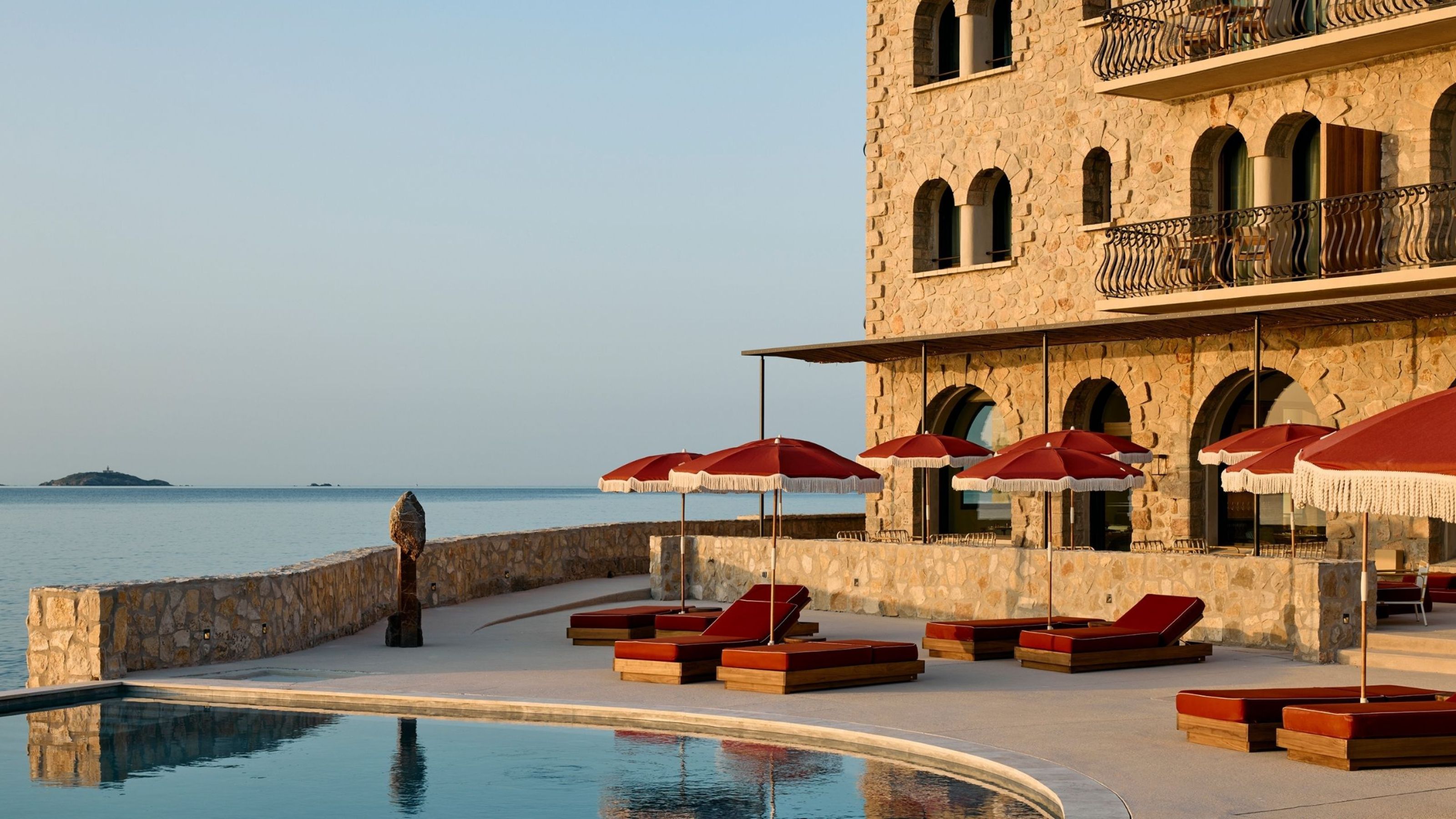 A sunset scene caught on the outdoor patio and pool of a hotel, decked out with dark red parasols and matching reclining loungers, an infinity pool, and uninterrupted sea views