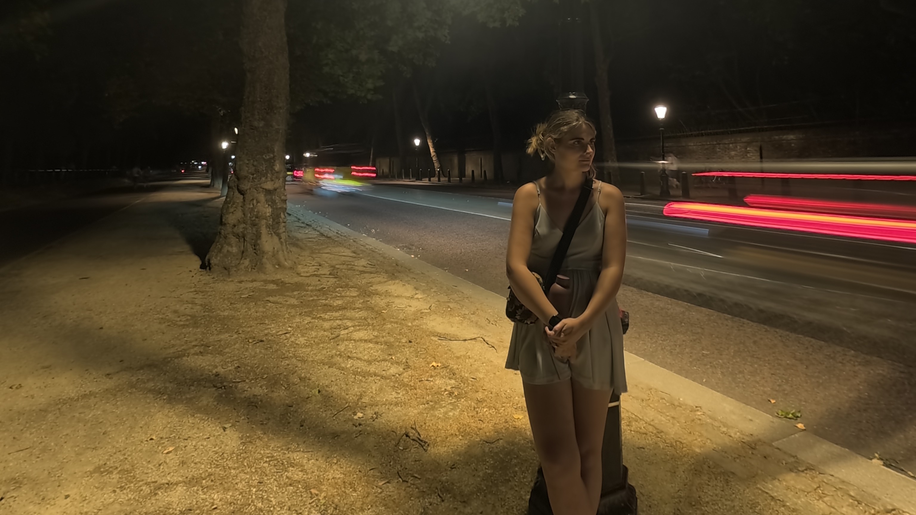 A woman standing by a lamp post at night as cars rush past