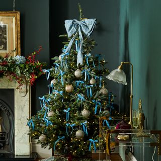 A dark green dining room with a Christmas tree decorated with blue ribbons, metallic baubles and an oversized blue bow as a tree topper