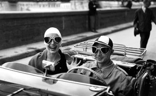 Black and white image of a couple in a convertible car with the rood down, photographer during the day with a bridge in the background