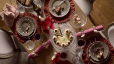 A table dressed in ruffled placemats ribbed plates, glass coupes, flutes, and a crystal cake stand with cake, a plate of macarons, by pink Christmas crackers