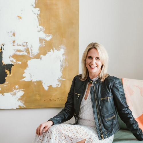 Image of a woman in a white dress and black leather jacket sitting in a white room with a large gold and white abstract painting on the wall.