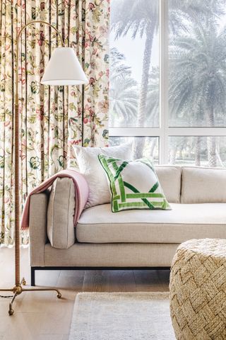 cream sofa with floral print curtain behind with view of palm trees outside and beige floor tiles and woven drum stool