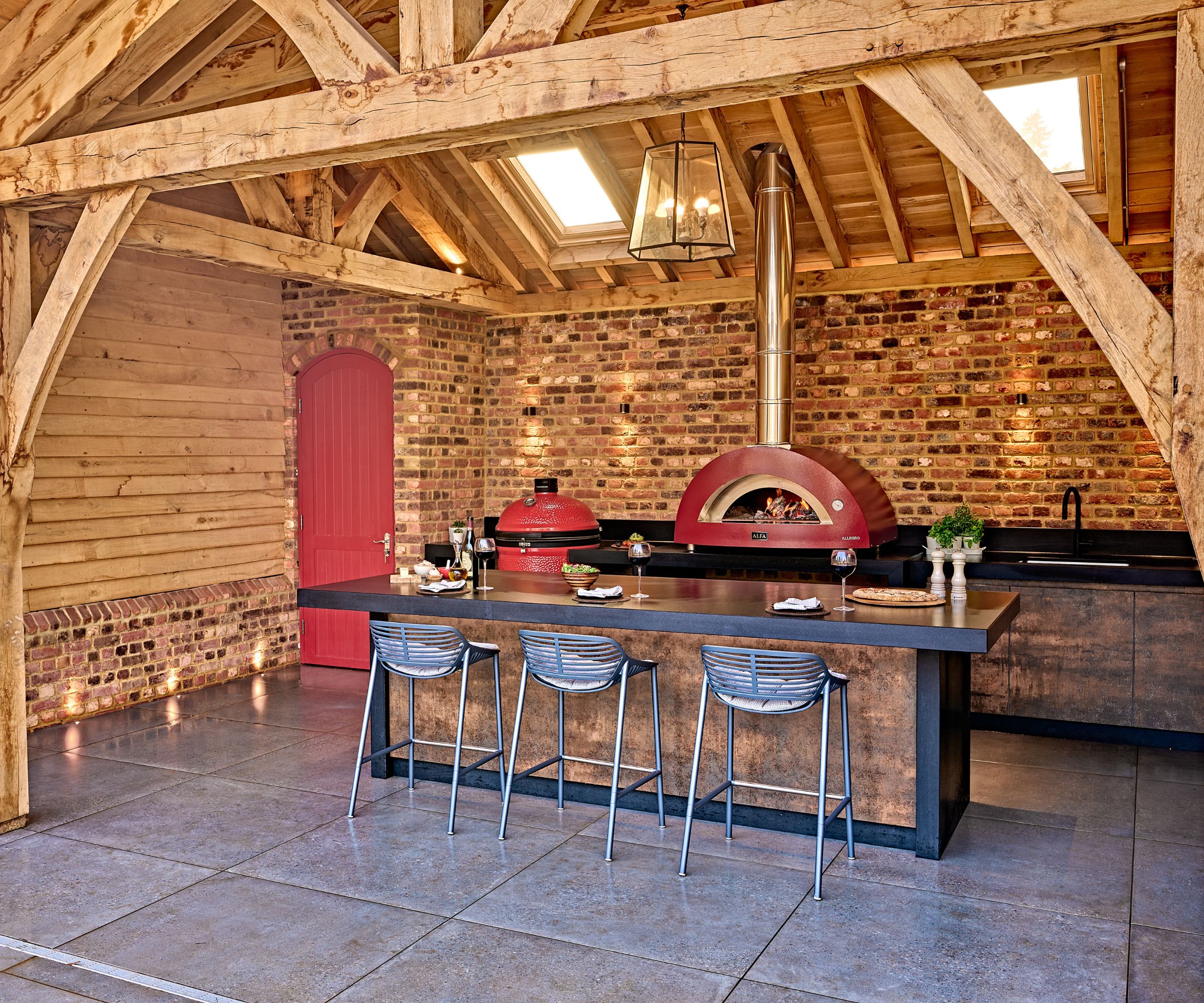 A covered outdoor kitchen with a red pizza oven, black countertops, and a wooden canopy