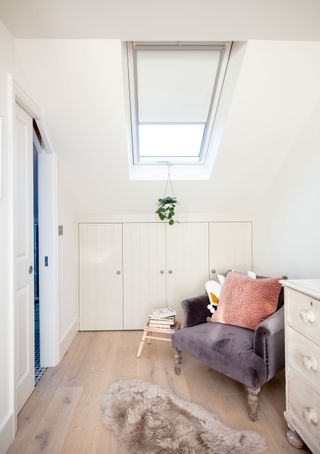 A white bedroom with wardrobe storage underneath eaves, grey velvet upholstered chair with pink cushion, white chest of drawers and small sheepskin rug decor