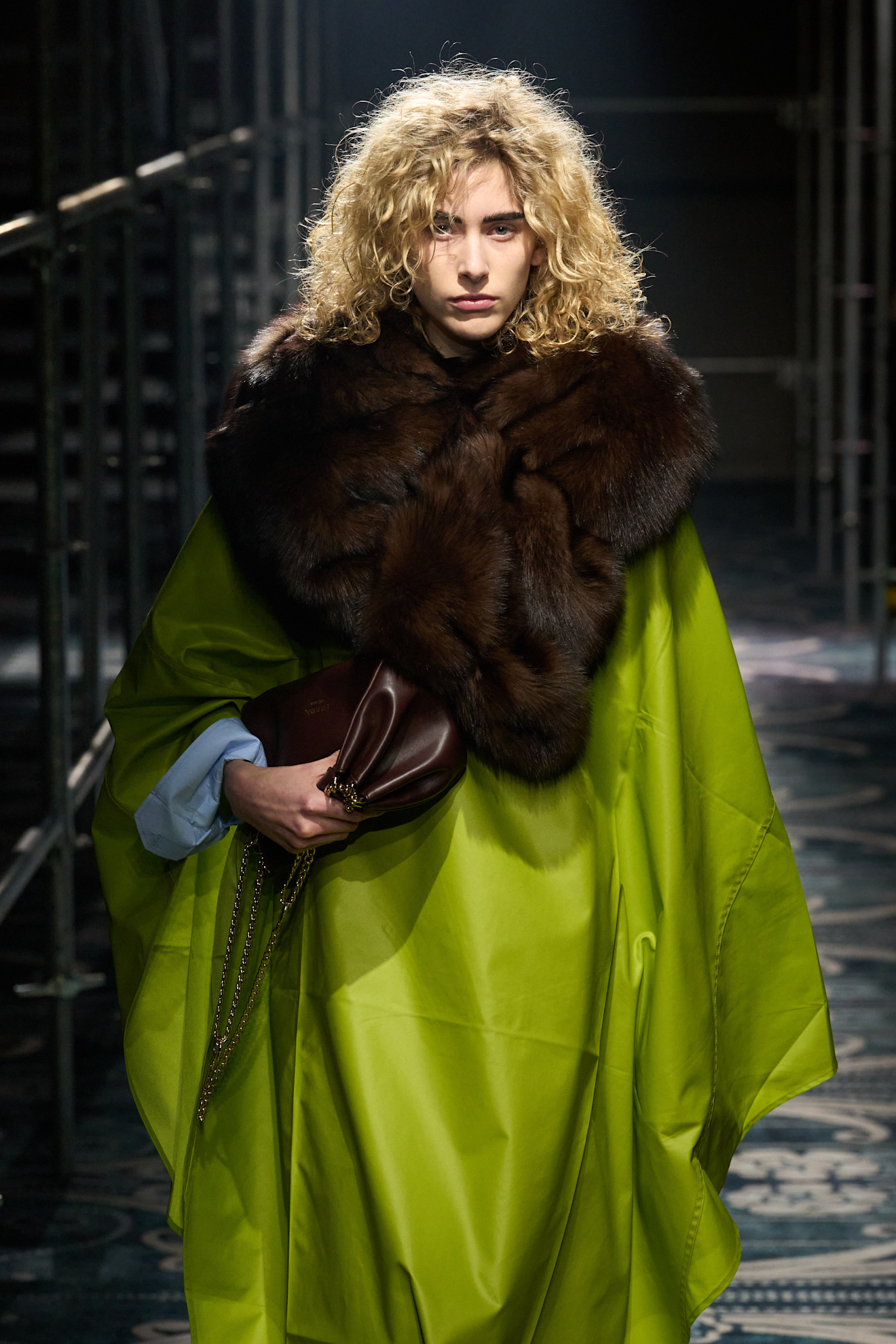 Fur accessory trend shown in Prada's fall-winter 2025 collection in a close photo of a model on the runway wearing a green silk cape with a dark brown fur stole around the neck while she carries a chocolate brown clutch bag