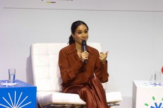 Meghan Markle sits onstage wearing a pumpkin spice orange shirt and matching pants