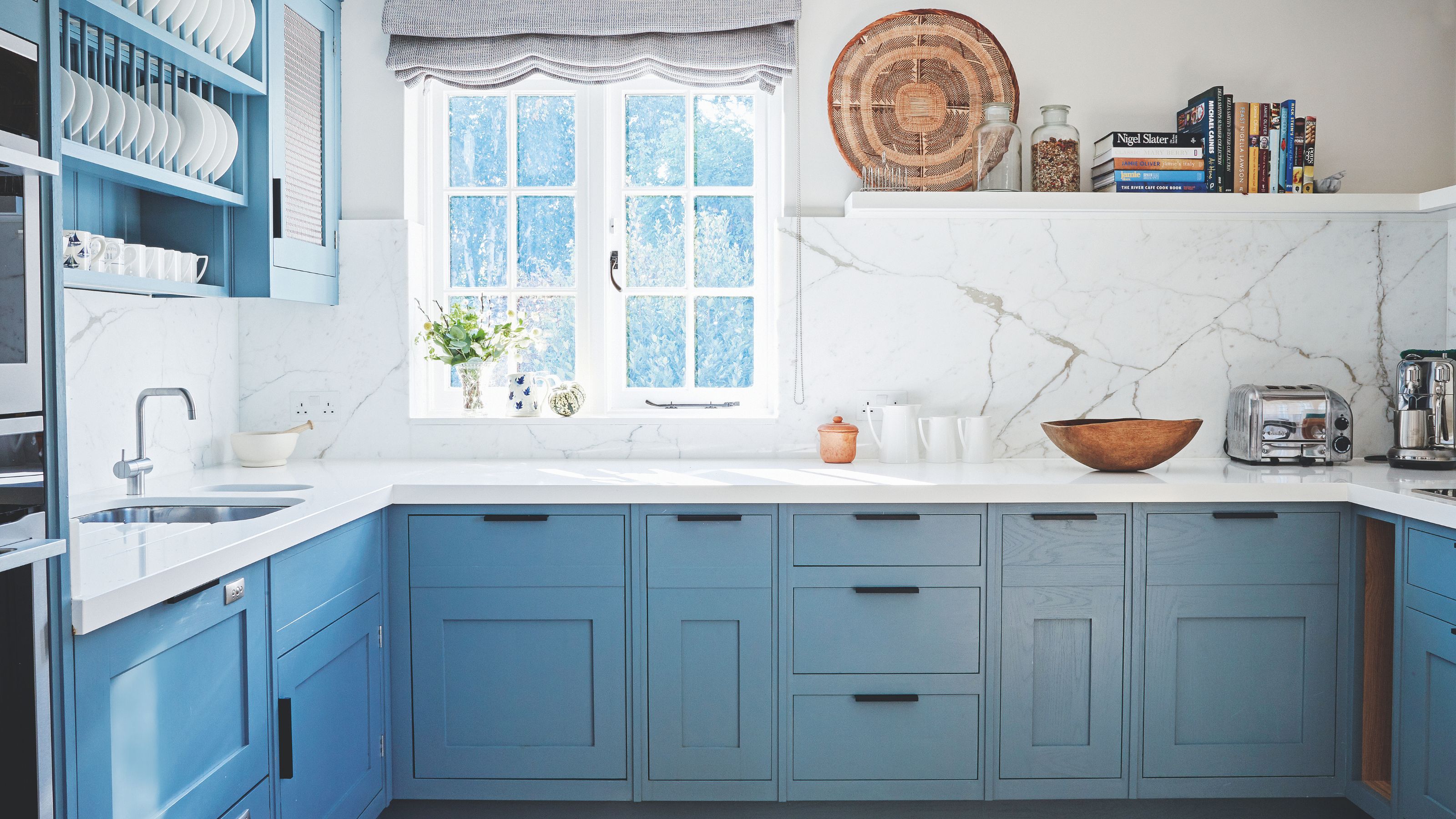White painted kitchen with white marble worktop and light blue cabinets underneath a window