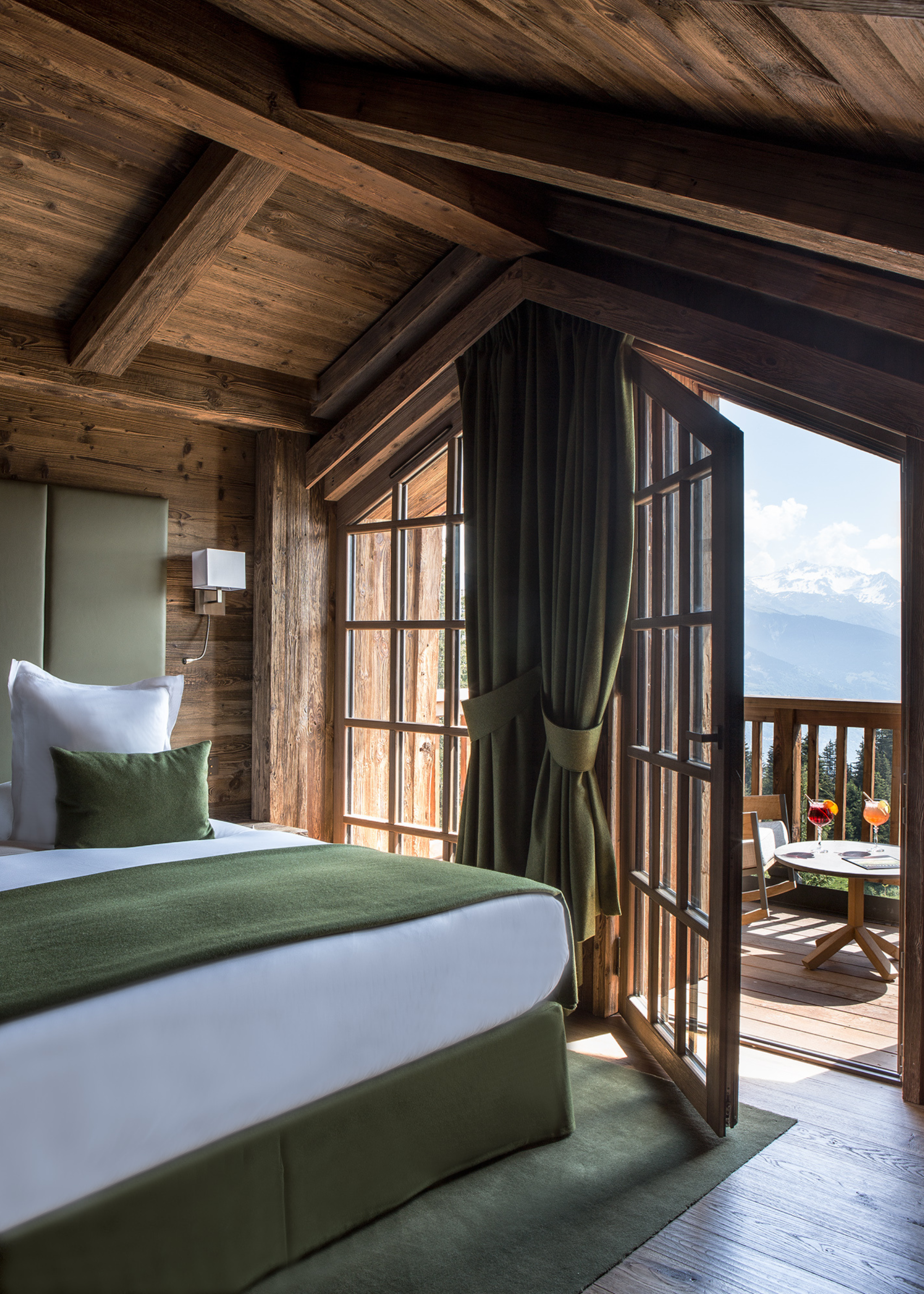 A chalet bedroom at Refuge de la Traye with a green color schemeand an open door leading onto the balcony with a table with cocktails on it to view the snowy mountains