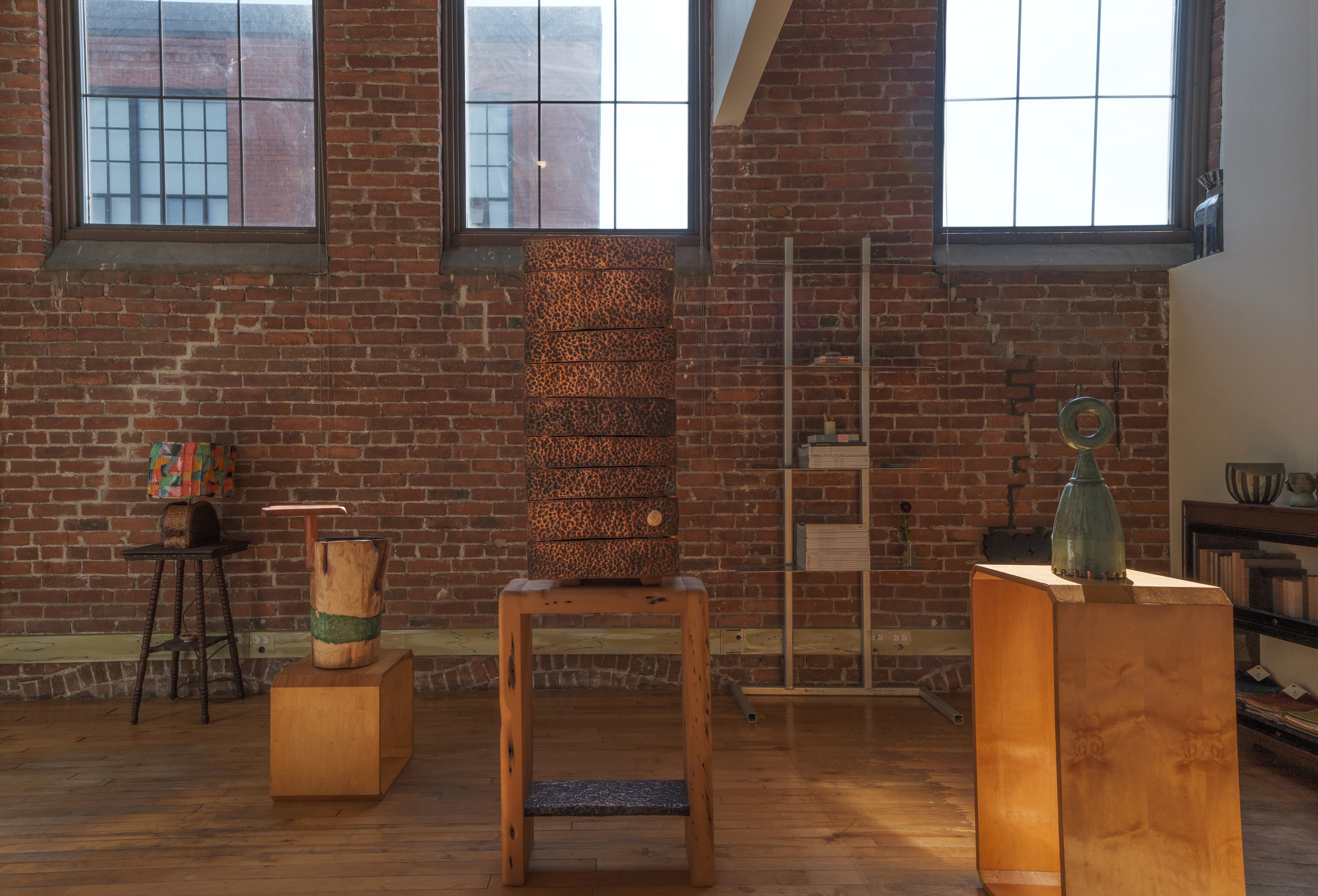 A gallery interior with a red-brick wall, and wood, ceramic, and bronze collectibles stacked atop wooden podiums.