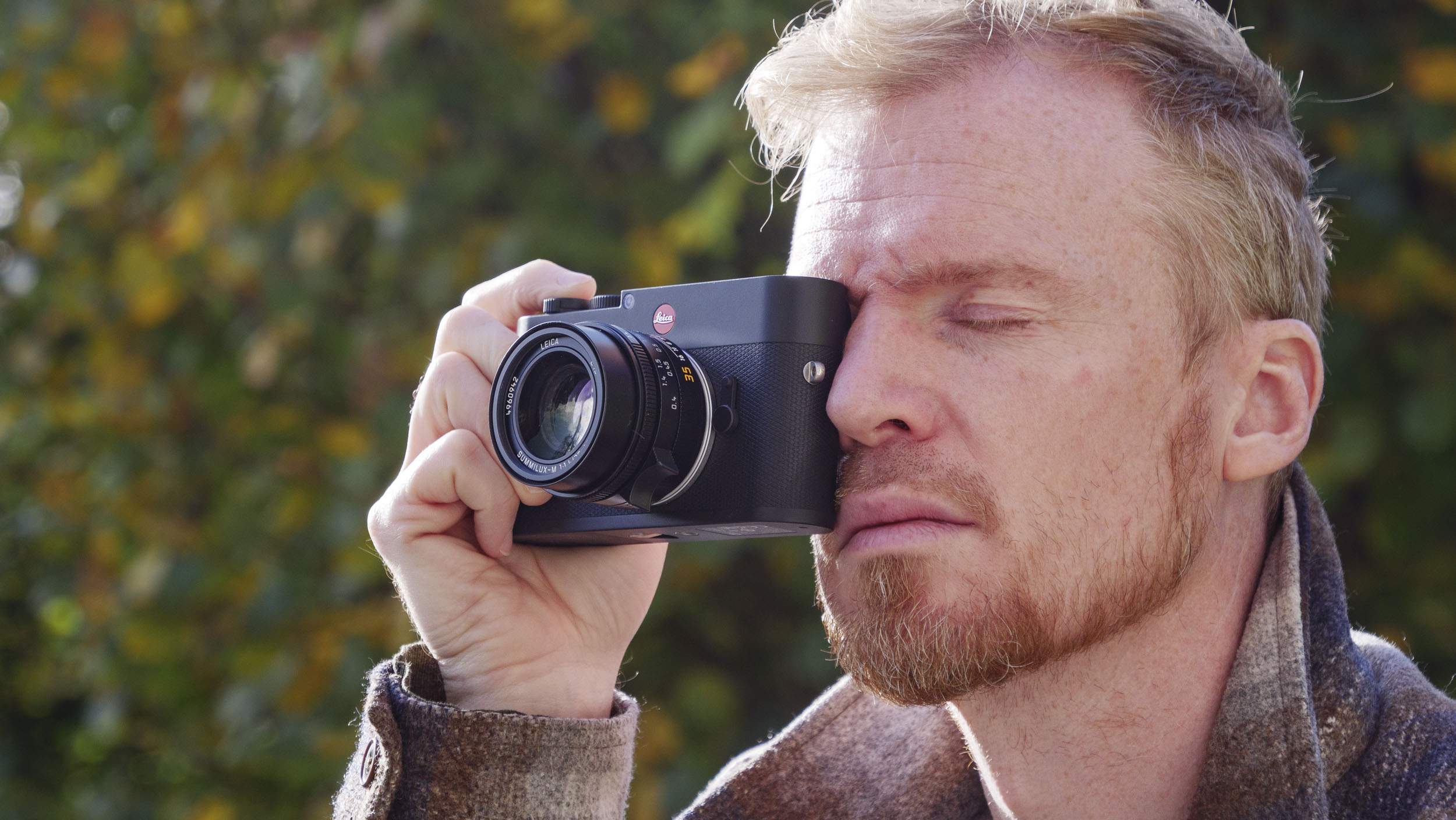 Man holding the Leica M EV1 camera's viewfinder up to their eye, autumn leaf backdrop