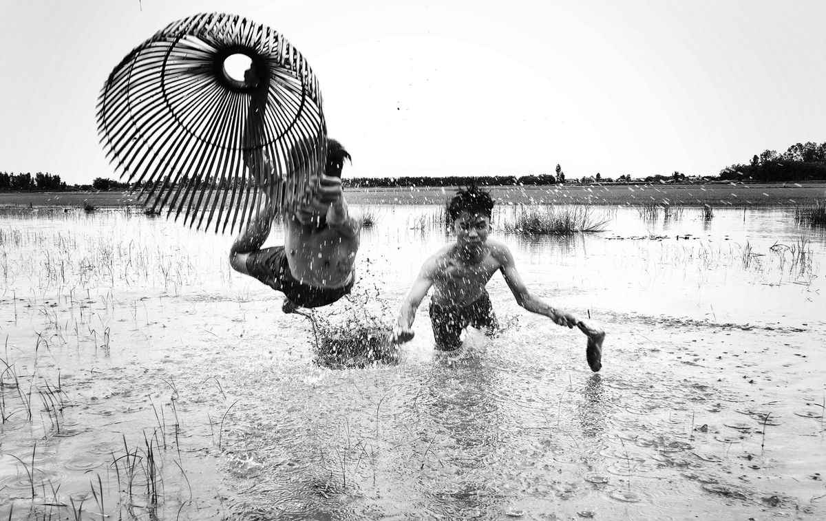 Two boys splash playfully in a muddy field, one jumping with a large woven fan, creating a dramatic spray of water around them
