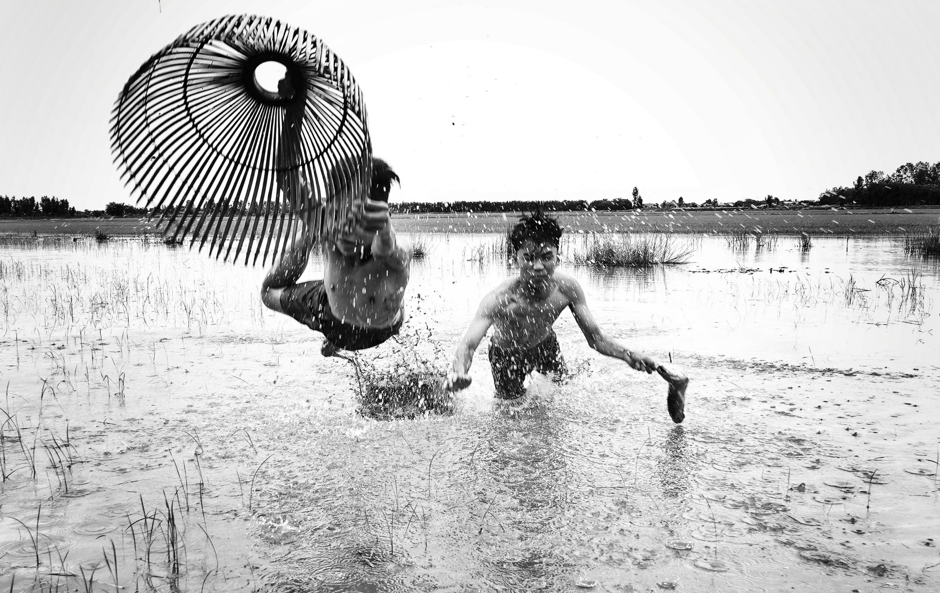 Two boys splash playfully in a muddy field, one jumping with a large woven fan, creating a dramatic spray of water around them