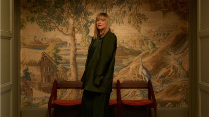 Image of interior designer Roisin Lafferty in an oversized suit standing in front of a mural wallpaper with a row of three red and wooden chairs.