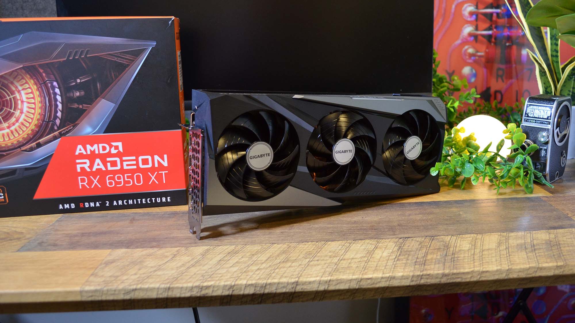 Ett AMD Radeon RX 6950 XT står uppställt på ett skrivbord bredvid sin tillhörande förpackning och en växt.