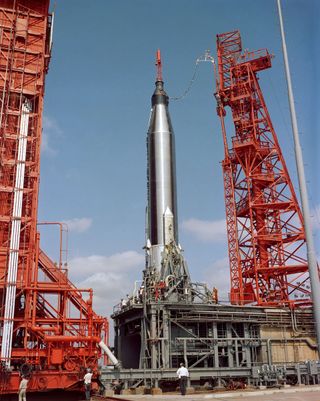 a silver rocket stands upright on a launch pad surrounded by two red metal towers