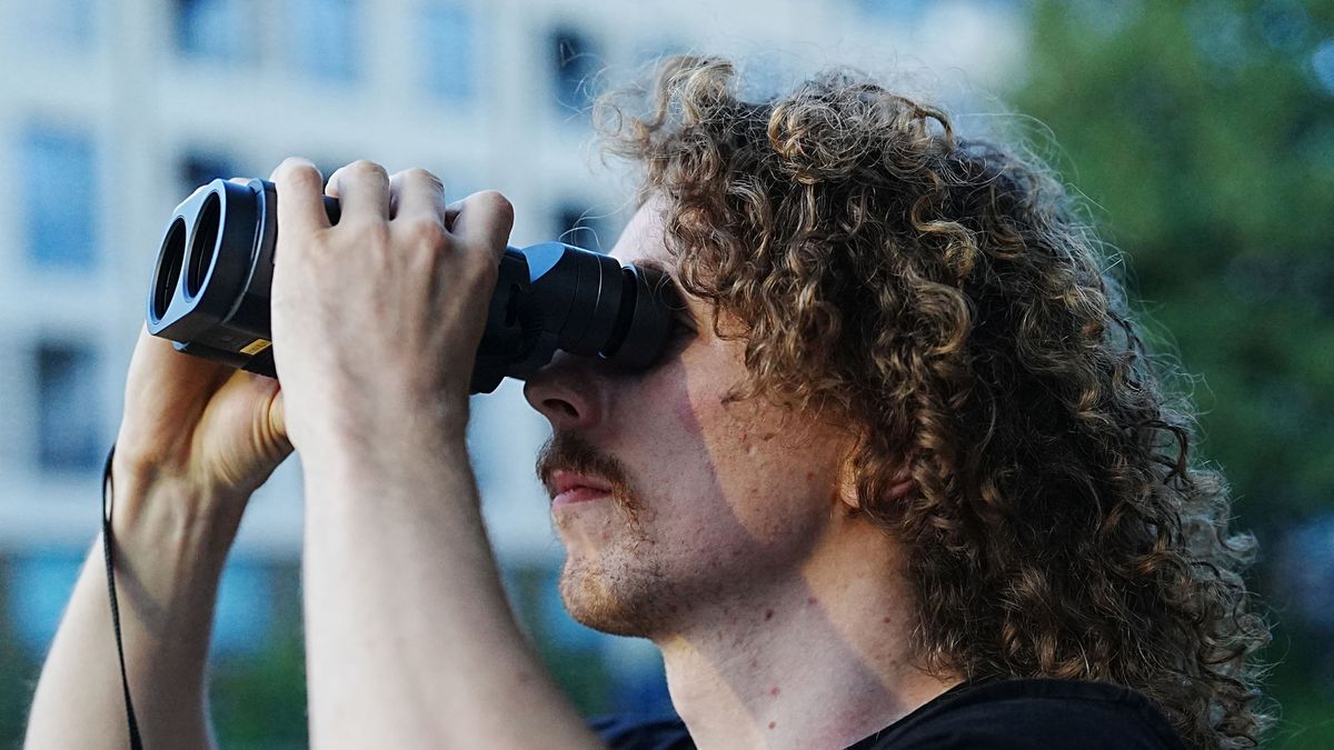 A close-up of a man looking through the Fujifilm Techno-Stabi TS-L 1640 image-stabilized binoculars with buildings and trees in the background.