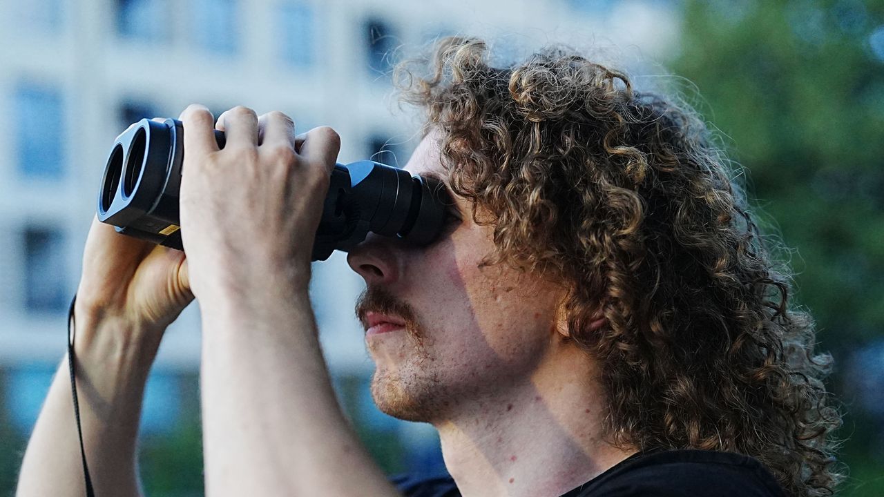 A close-up of a man looking through the Fujifilm Techno-Stabi TS-L 1640 image-stabilized binoculars with buildings and trees in the background.