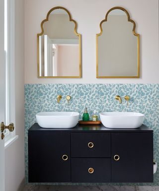 A bathroom with two gold arched mirrors on the wall, a blue and white flowery splashback, and two dual sinks with white basins and a navy blue rectangular base