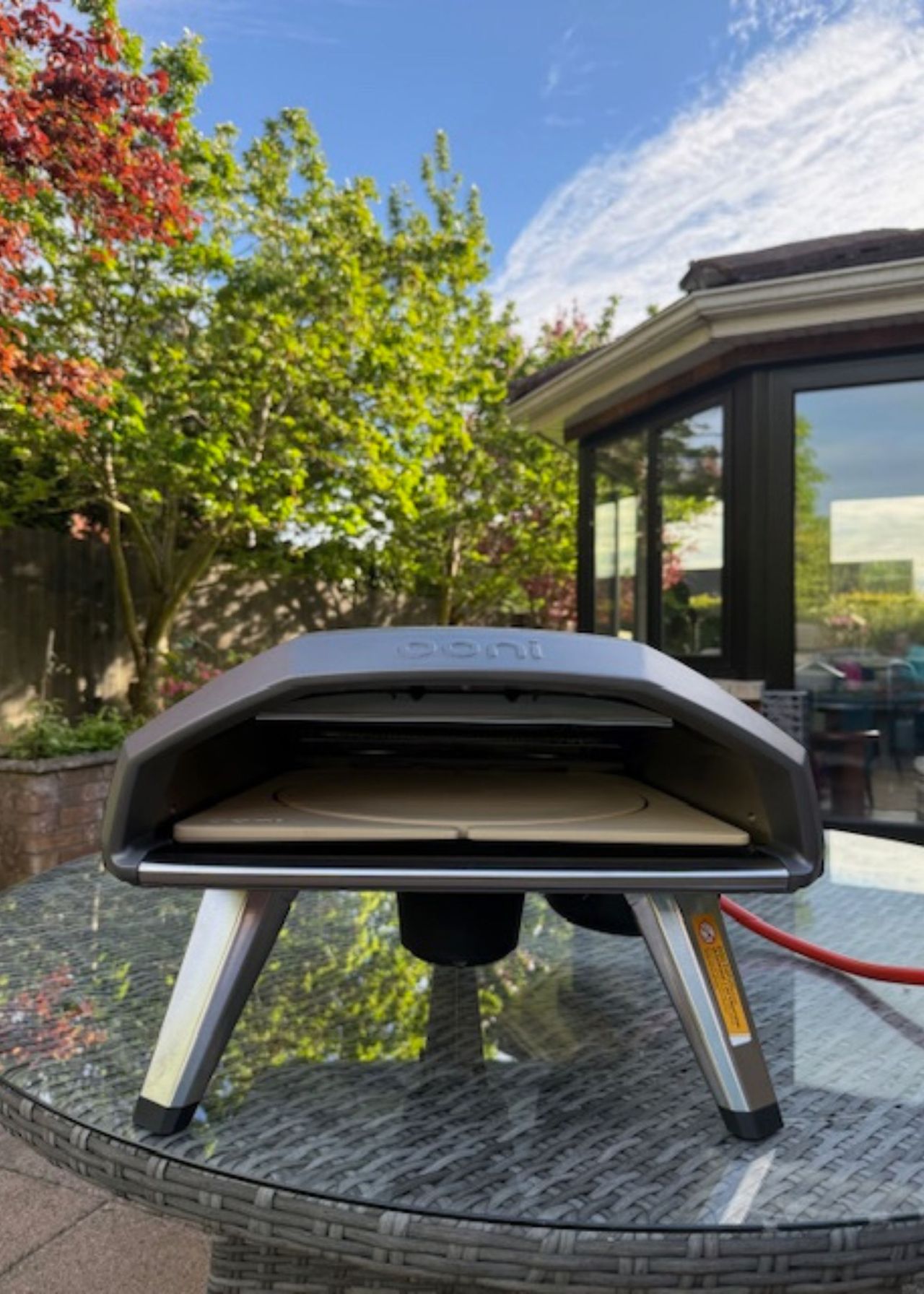 Ooni Koda 2 Rotating Stone on a table in a garden with a sunroom with black-framed windows in the background next to green and red-leaved trees below a blue sky